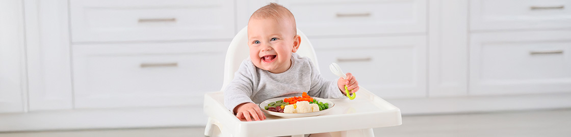 foto de un bebé feliz sentado en su silla de bebé y comiendo sus primeros alimentos sólidos (puré y verduras)