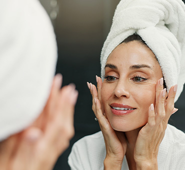 Foto de mujer en toalla frente al espejo del baño aplicando cremas de skincare en su rostro.