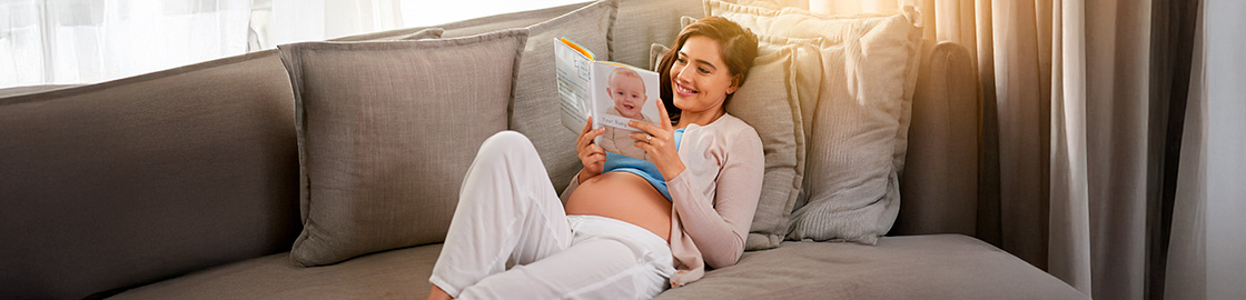 Foto de mujer embarazada acostada en el sillón leyendo un libro sobre maternidad
