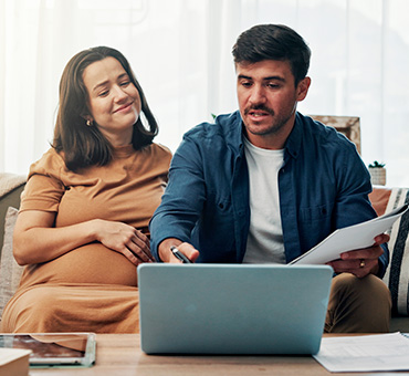 Foto de mujer embarazada sentada junto a su pareja mientras ven algo en la laptop