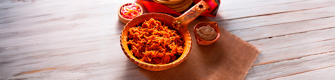 Foto de una cazuela de cerámica con cochinita pibil, servida junto a frijoles y cebolla morada