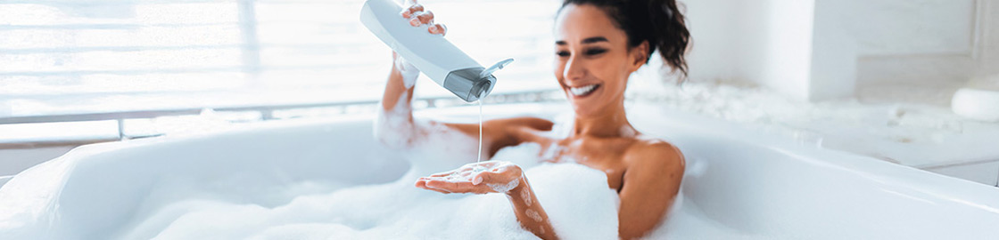 Foto de mujer bañándose en una tina, cubierta de espuma y vertiendo shampoo en su mano mientras sonríe.