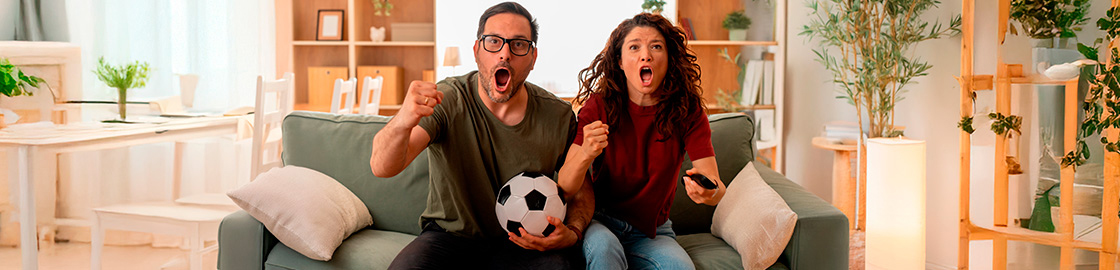 Foto de una pareja sentada en el sillón, celebrando un gol mientras el chico sostiene un balón de fútbol.