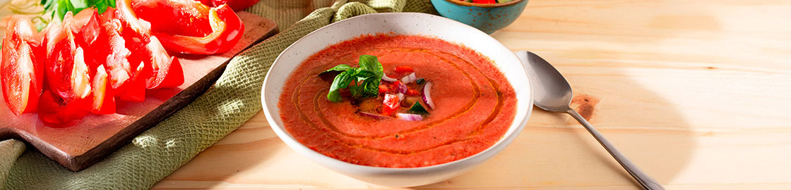 Foto de un plato de gazpacho servido sobre una mesa de madera, junto a una cuchara, con jitomates y pimientos de fondo.