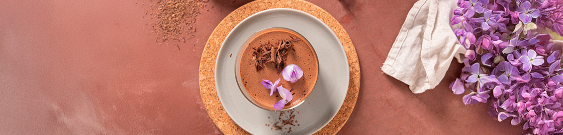Foto de una copa con mousse de chocolate, decorado con pétalos de flores, servido sobre un plato en una mesa rústica.