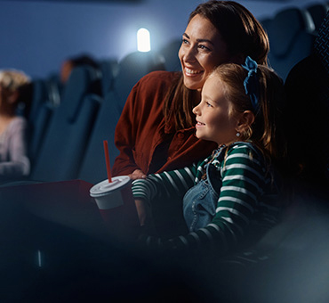 Foto de una niña contenta sentada en el cine viendo la pantalla con su mamá al lado.