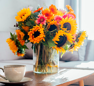 Foto de un florero lleno de girasoles en la mesa de una sala de estar, al lado de una taza de café.
