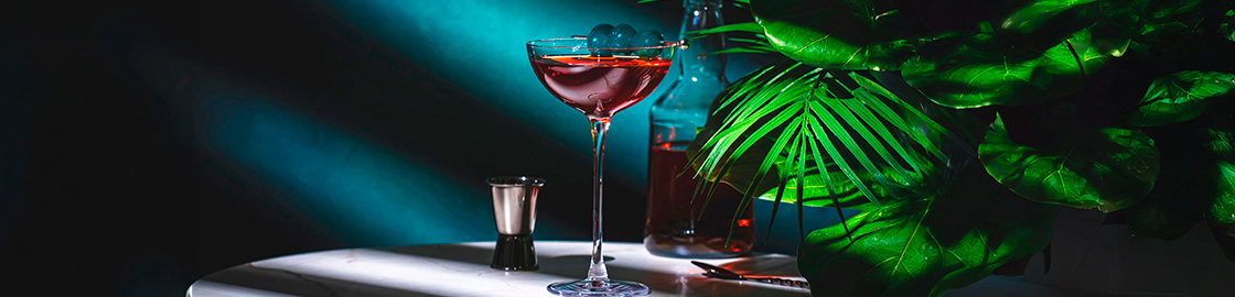 Foto de un coctel Rob Roy junto a una botella de whisky y vermut sobre una mesa con fondo de pared azul y plantas decorativas junto a las bebidas