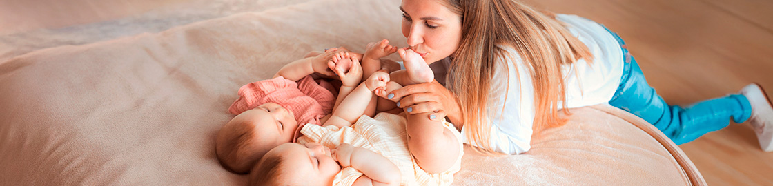 Foto de dos bebés gemelos acostados en la cama y su mamá, agachada frente a ellos, besando sus pies
